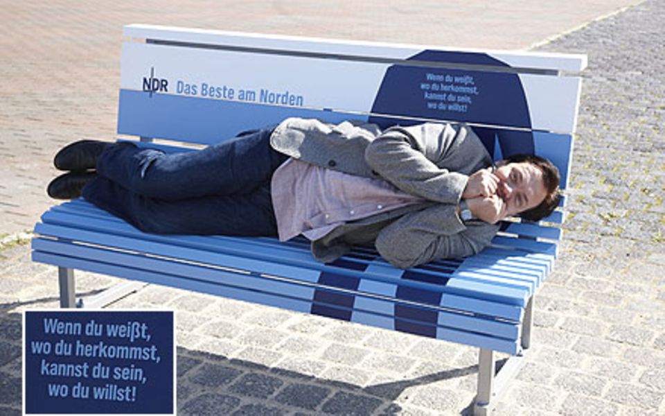 Schauspieler Charly Hübner macht auf seiner Bank im Stadthafen von Rostock Faxen.