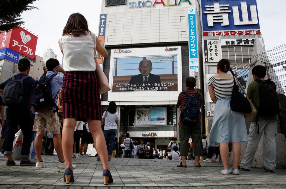 In Tokio stoppen Menschen im Vorbeigehen und hören dem Kaiser und seiner Ansprache zu.