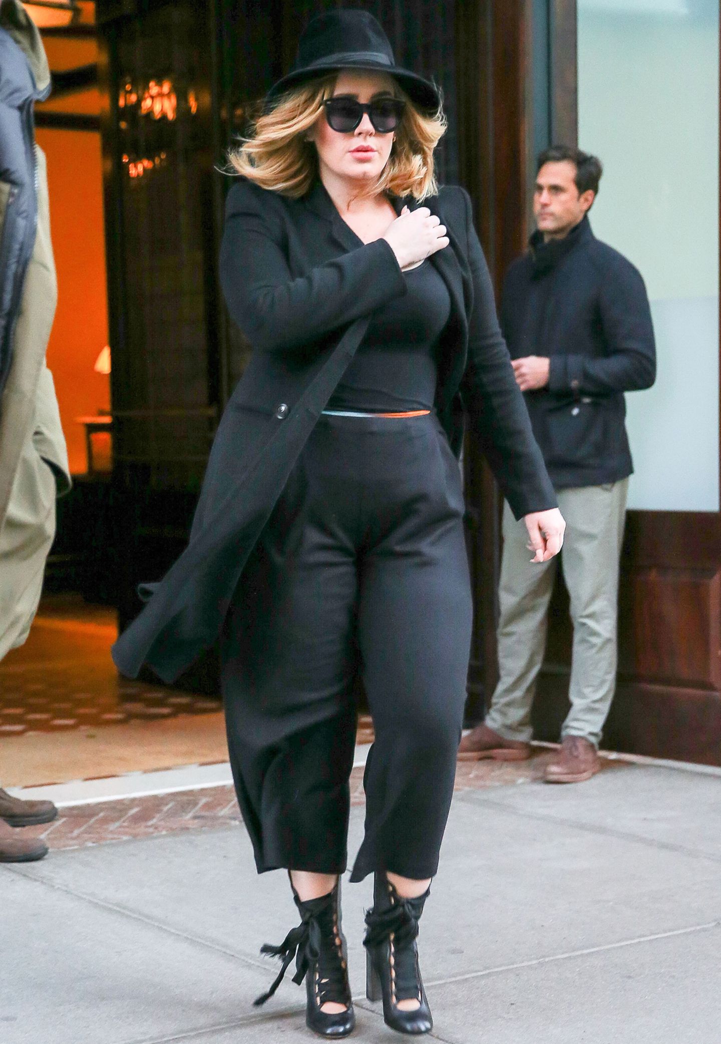 Schwarze Culottes, Stilettos mit Schnürung, taillierter, schwarzer Mantel, Sonnenbrille und Hut - Adele sieht aus wie ein echter Megastar.