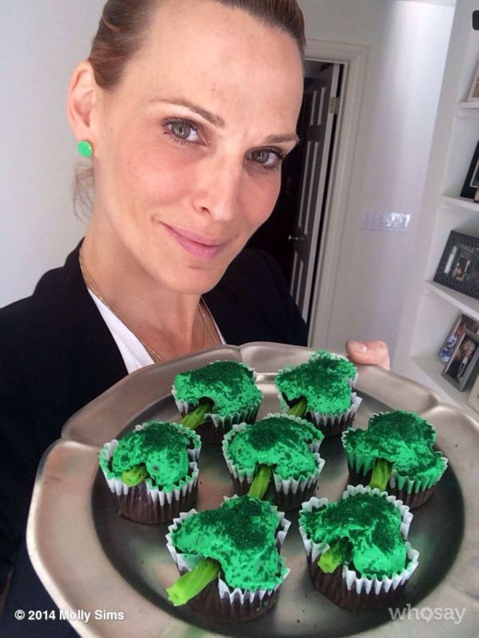 Molly Sims posiert mit grünen Ohrringen und Cupcakes in Kleeblatt-Form für ein Foto.