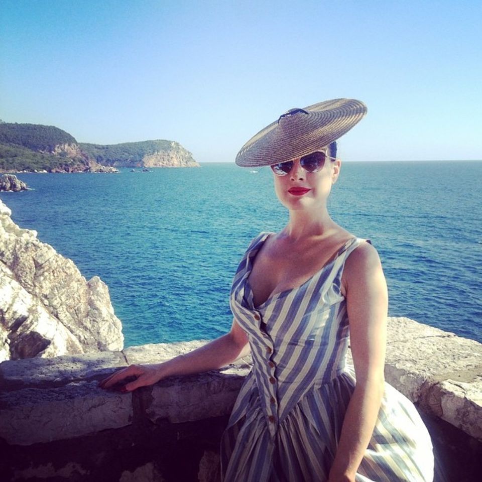 Die sonst so burlesque Dita von Teese wirkt auf ihren Instagram-Urlaubsgrüßen wie auf einer Postkarte aus den Fifties: Tellerstrohhut mit gestreiftem und geknöpftem Sommerkleid passen toll zum Strand-Look.