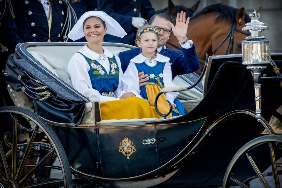 Prinzessin Estelle und Mama Victoria präsentieren sich in der blau-gelben Nationaltracht. Prinz Daniel winkt dem Volk zu. 