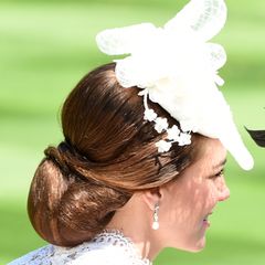 Auf dem Kopf trägt sie einen weißen Faszinator mit Blumendekoration. Ihr brünettes Haar ist zu einem eleganten Chignon zusammen geknotet. 