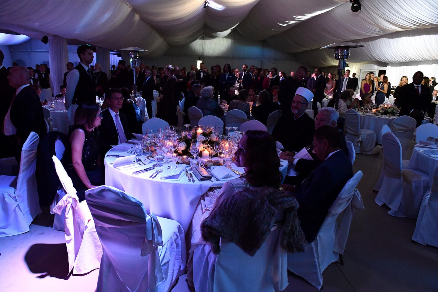 Die Gäste sitzen während der Feier an großen runden Tischen, der Raum ist mit einem Baldachin ausgekleidet und in blaues Licht getaucht. Die romantische Atmosphäre entsteht durch Kerzenschein  und Rosengedecke.