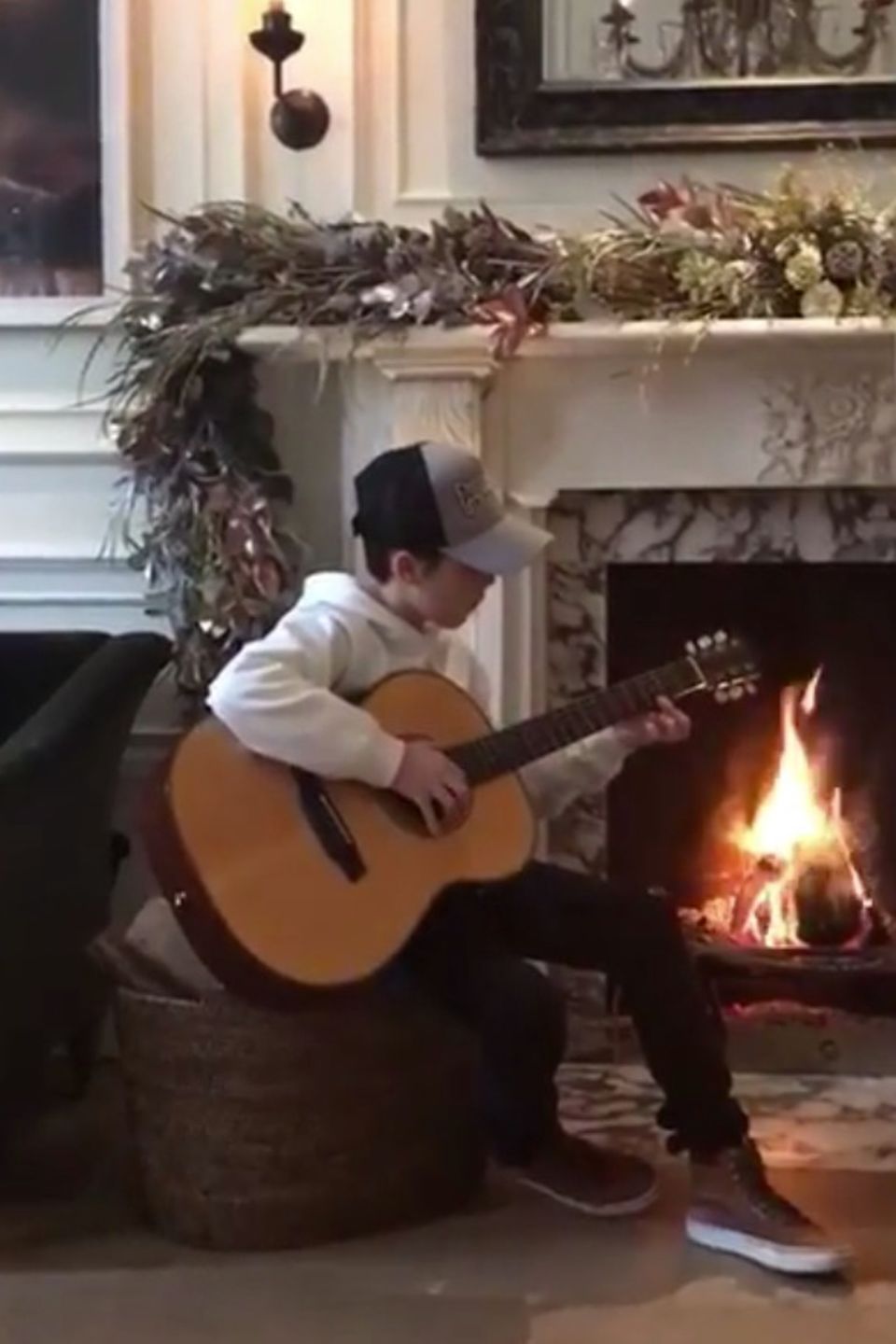 Der musikalische Cruz Beckham kommt ganz nach der Mama Victoria. Vor dem Karmin im heimischen Wohnzimmer übt der Sprössling Gitarre. Der Karmin aus Marmor ist mit einer opulenten Blumendekoration versehen.