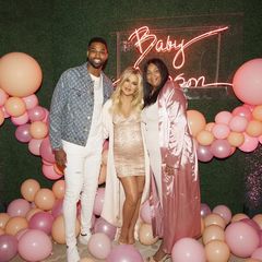 Die hochschwangere Khloé Kardashian posiert mit ihrem Freund und Bald-Papa Tristan Thomson und dessen Mutter Andrea Thompson. Khloé trägt ein hellrosa Kleid mit Glitzerapplikationen, dass sich um ihren Babybauch schmiegt und kombiniert dazu einen passenden cremefarbenen Mantel.
