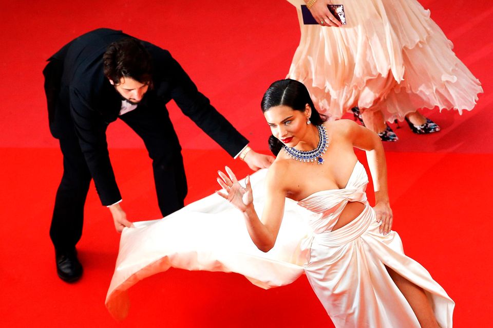 Supermodel Adriana Lima winkt entspannt den Besuchern zu, während der fleißige Assistent mit ihrer wallenden Schleppe zu kämpfen hat.