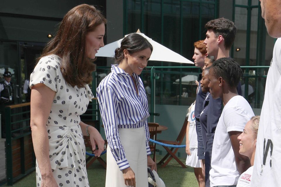 In einer weißen Marlenehose und einer blau-weiß-gestreiften Bluse von Ralph Lauren erscheint Herzogin Meghan an der Seite ihrer Schwägerin. Gemeinsam begrüßen sie vor dem Spiel die Balljungen und -Mädchen. 
