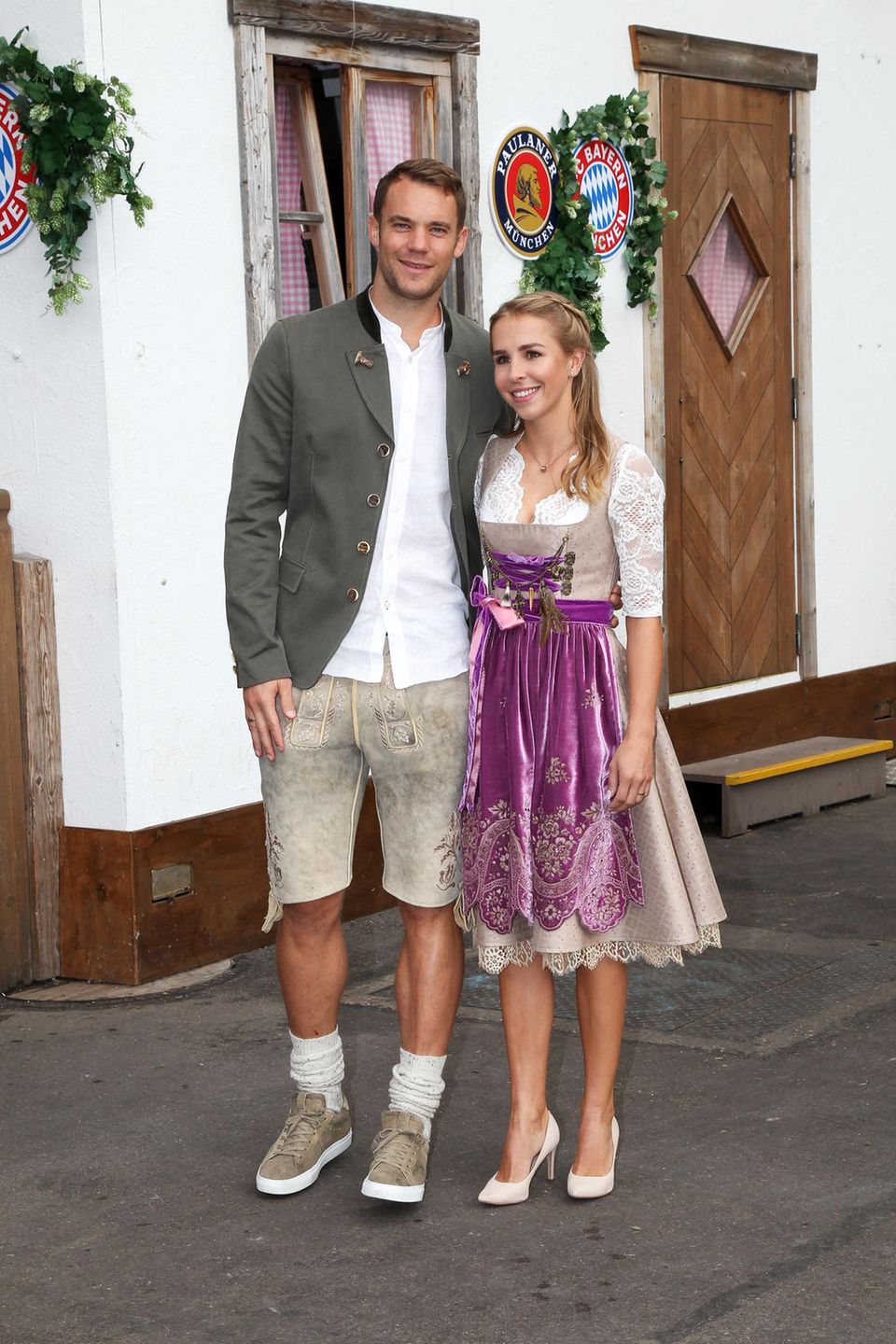 Auch Torwart Manuel Neuer und seine Frau Nina dürfen bei dem Besuch des FC Bayern Münchens auf der Wiesn nicht fehlen. Das Paar hat seine Looks farblich aufeinander abgestimmt. Lisa Neuer trägt ein beigefarbenes, edles Dirndl mit lilafarbener Samt-Schürze, Manuel trägt eine Lederhose in einem ähnlichen Beige-Ton. 