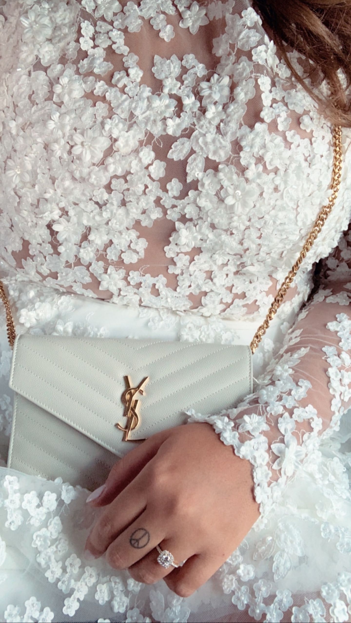 Das Kleid aus dem Düsseldorfer Brautmodengeschäft "IamYours" verzaubert mit durchsichtigen Einsätzen, die von vielen kleinen, weißen Blüten geziert werden. Dazu kombiniert sie eine kleine weiße Tasche von Saint Laurent. 