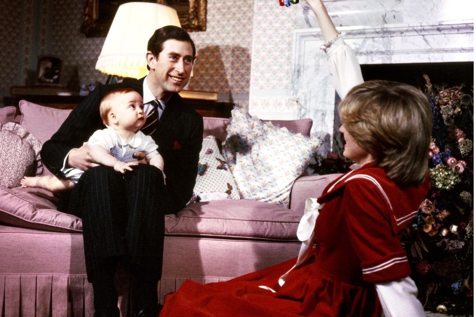 1982  Der kleine Prinz William sitzt auf dem Schoß seines Vaters Prinz Charles und schaut fasziniert auf das Spielzeug, das seine Mutter Prinzessin Diana hochhält.