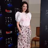Katie Holmes posiert in einem weißen Blüschen und in einem gemusterten, knielangen Rock auf dem roten Teppich. Mit roten Lippen und blauen Heels setzt sie zusätzlich farbliche Akzente - doch das sind nicht die einzigen... 
