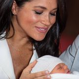 Herzogin Meghan + Prinz Harry: Meghan hält ihre Hand schützend an den Kopf ihres Babys, das eine leichte Mütze trägt und in eine weiße Decke gehüllt ist.