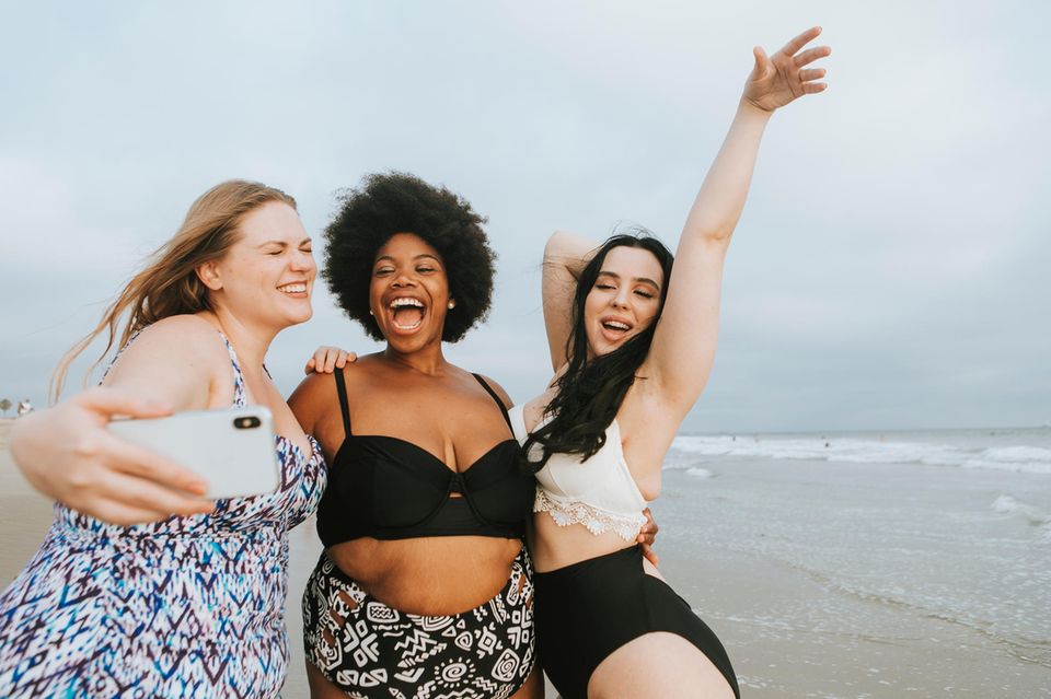 Shape Bikini, drei lachende Frauen in Bademode am Strand, die ein Selfie machen