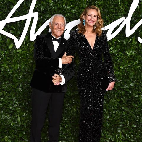 Hollywood-Glamour in London: Eine strahlende Julia Roberts erscheint Hand in Hand mit Designer Giorgio Armani. Sie trägt eine schwarze Glitzerrobe mit tiefem V-Ausschnitt und hat ihr Haar in elegante Wellen gelegt. 