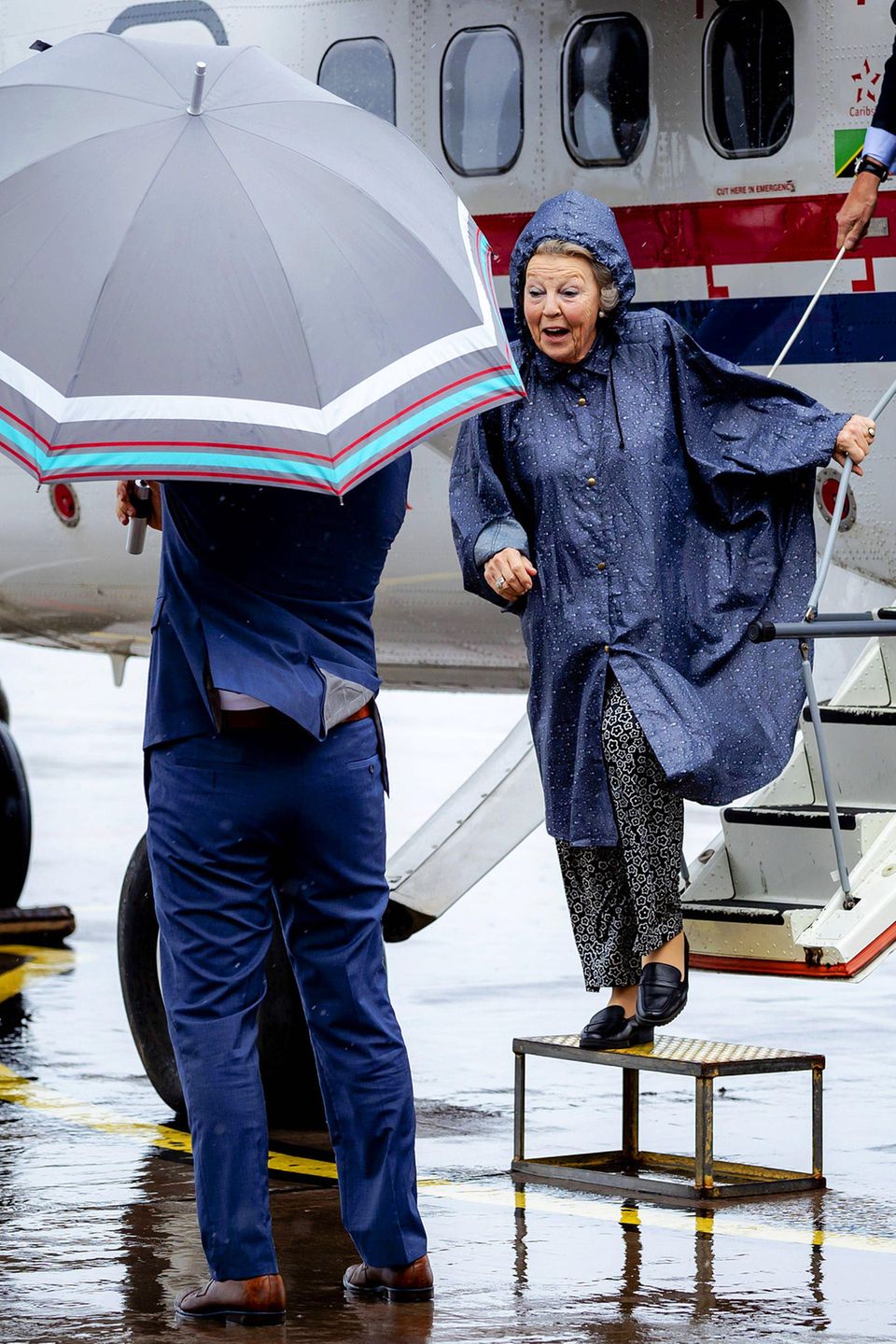 Bei der Ankunft von Prinzessin Beatrix auf der Insel Saba regnet es in Strömen. Zum Glück ist die Prinzessin mit einem Regenmantel gegen das schlechte Wetter bestens gerüstet und auch ein Schirmträger steht schon bereit. 