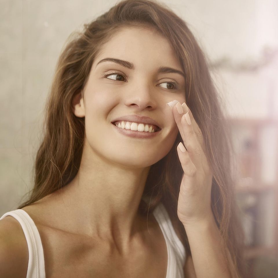 Schöne Frau mit braunen Haaren trägt Creme vor dem Spiegel auf ihr Gesicht auf.