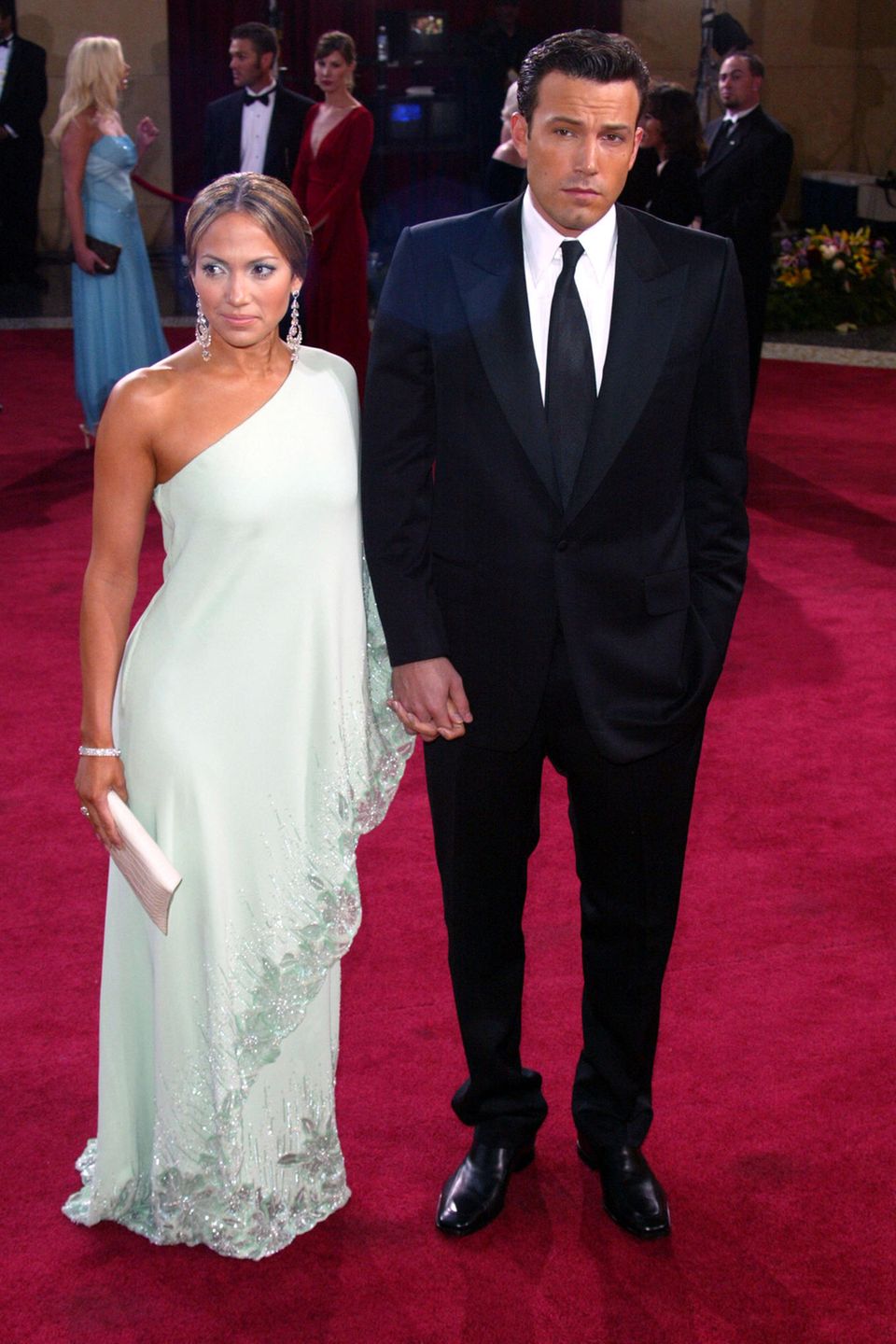 Beinahe schüchtern wirken Jennifer Lopez und Ben Affleck nachdem sie den Red-Carpet der Oscars im Jahr 2003 betreten haben. J.Lo trägt ein One-Shoulder-Dress mit Glitzer-Applikationen am Saum in der Farbe Weiß. Ben trägt einen schwarzen Anzug mit Krawatte. Unsichere Blicke, die Hände ineinander verankert. 