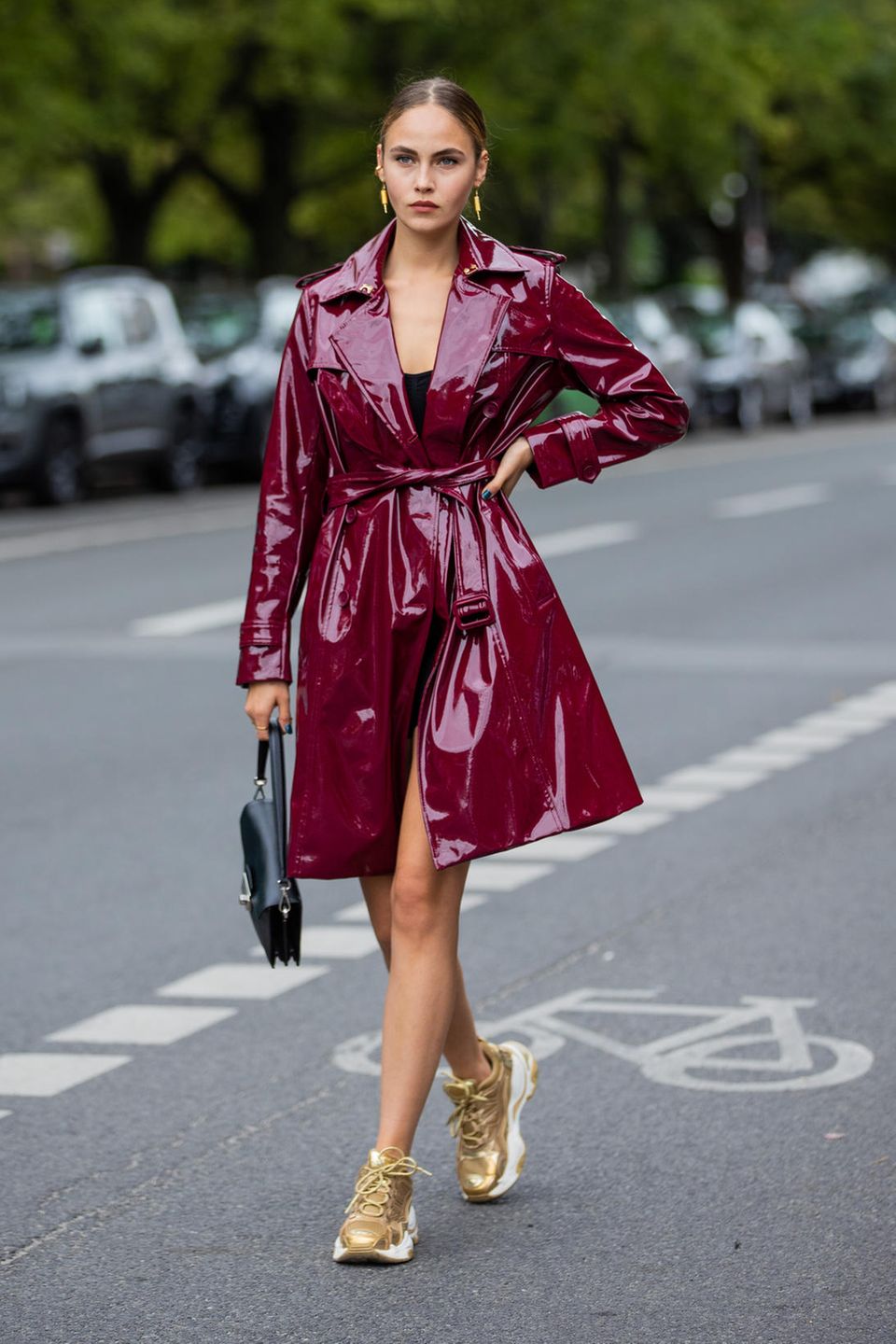 Model und Schauspielerin Elena Carriere trägt einen burgunderfarbenen Trenchcoat in Lack-Optik. Zu dem ausgefallenen Mantel wählt sie goldene Sneaker und macht mit der Kombination den Streetstyle-Look perfekt.