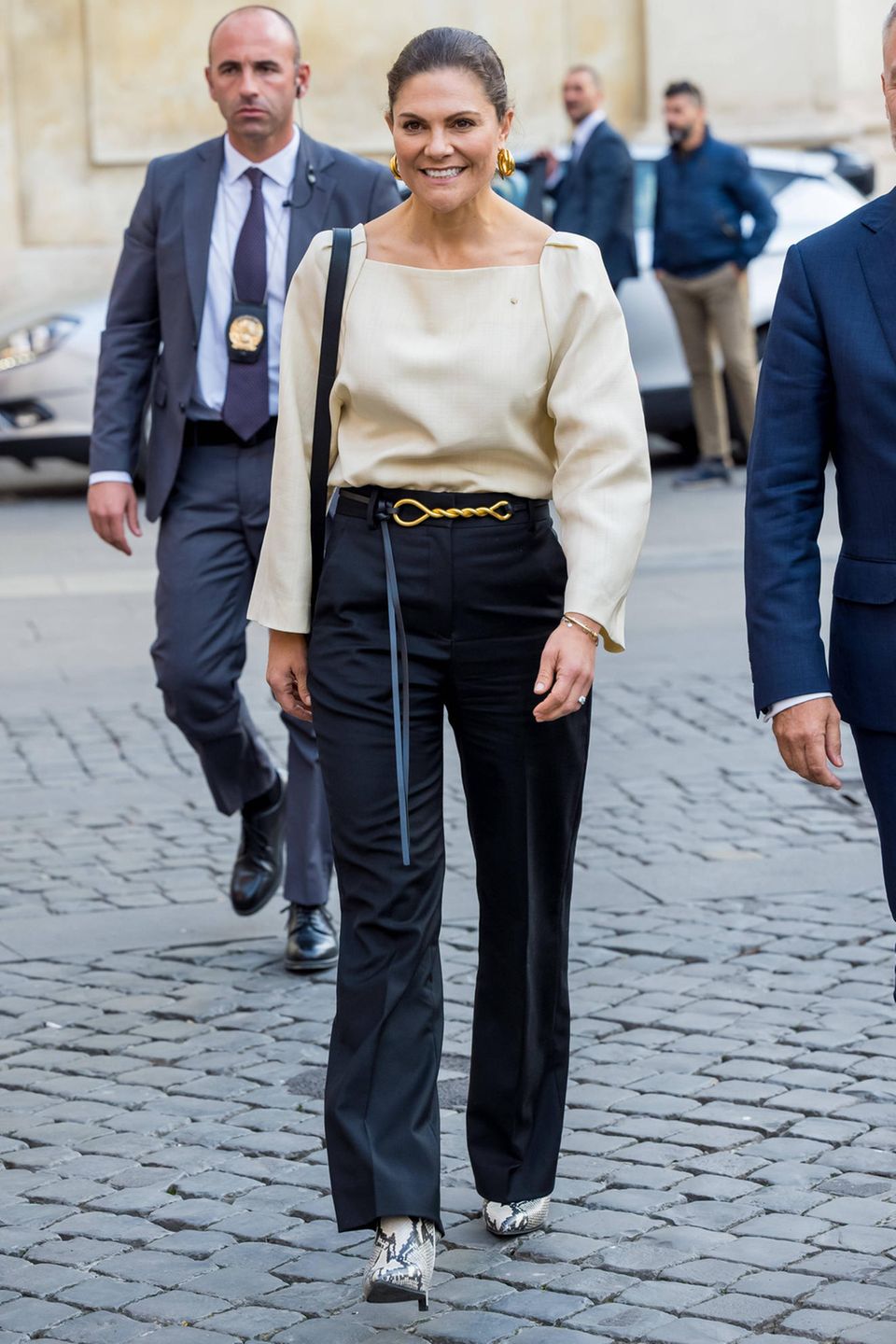 Bei dem Besuch einer Bilderausstellung in Italien zeigt sich Prinzessin Victoria in einem eleganten Look aus cremefarbener Bluse und dunkelblauer Anzugshose mit goldenem Gürtel. Das schlicht anmutende Outfit besticht durch eine überraschende Schuhwahl: Die Ehefrau von Prinz Daniel trägt auffällige Ankle Boots in Schlangenleder-Optik des schwedischen Labels Stinaa.J, die dem Look eine trendige Note verleihen.