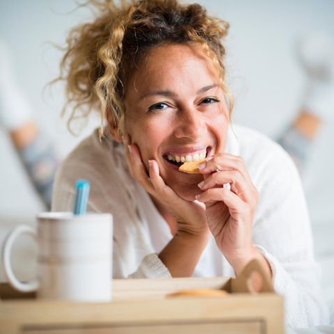 Horoskop: Lachende auf dem Bett liegende Frau isst einen Keks