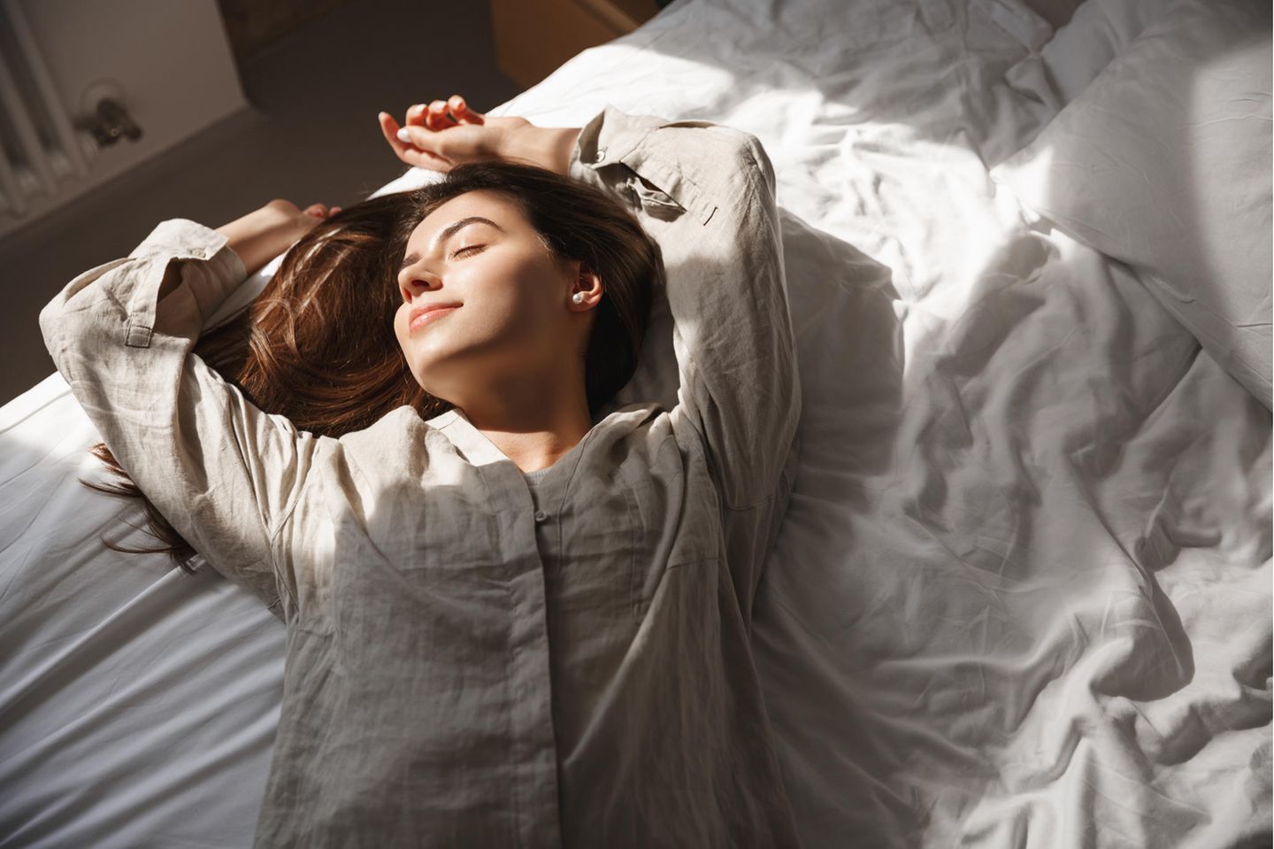 Brünette Frau liegt im Leinen-Pyjama auf Bett mit weißen Laken, Sonne strahlt ihr ins Gesicht