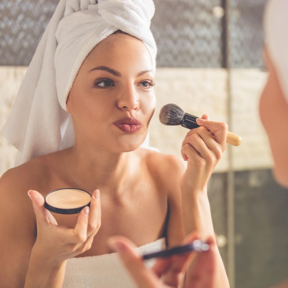 Eine Frau guckt lächelnd in den Spiegel und schminkt sich mit einem Make-up-Pinsel