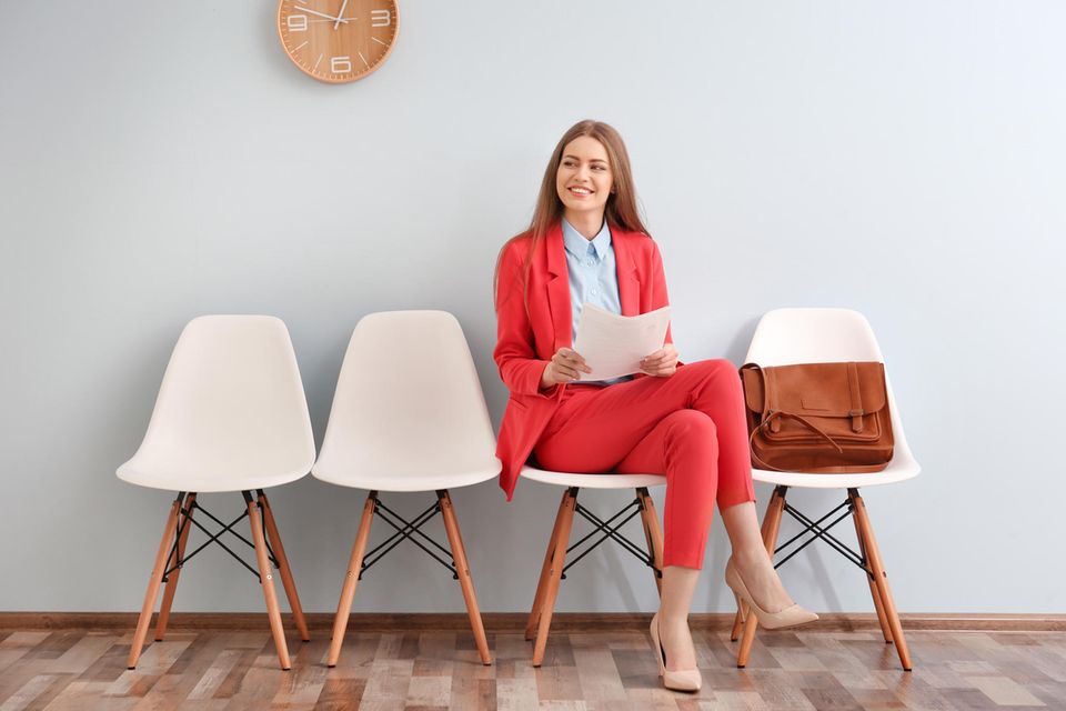 Eine blonde Frau trägt einen roten Anzug, sitzt auf einem Stuhl und wartet auf ihr Bewerbungsgespräch