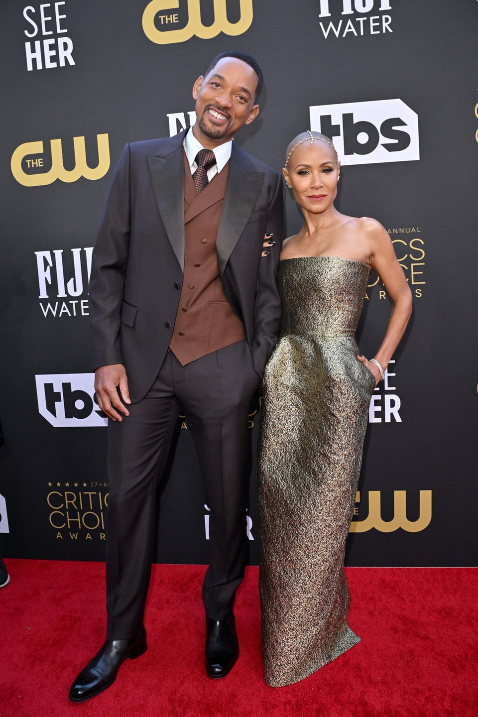 Gemeinsam posieren Will Smith und seine Frau Jada Pinkett Smith auf dem Red Carpet. Will setzt auf eine Kombi aus Schwarz und Braun; Jada dagegen auf ein gold-graues Kleid ohne Träger. Dazu trägt sie goldenen Kopfschmuck.