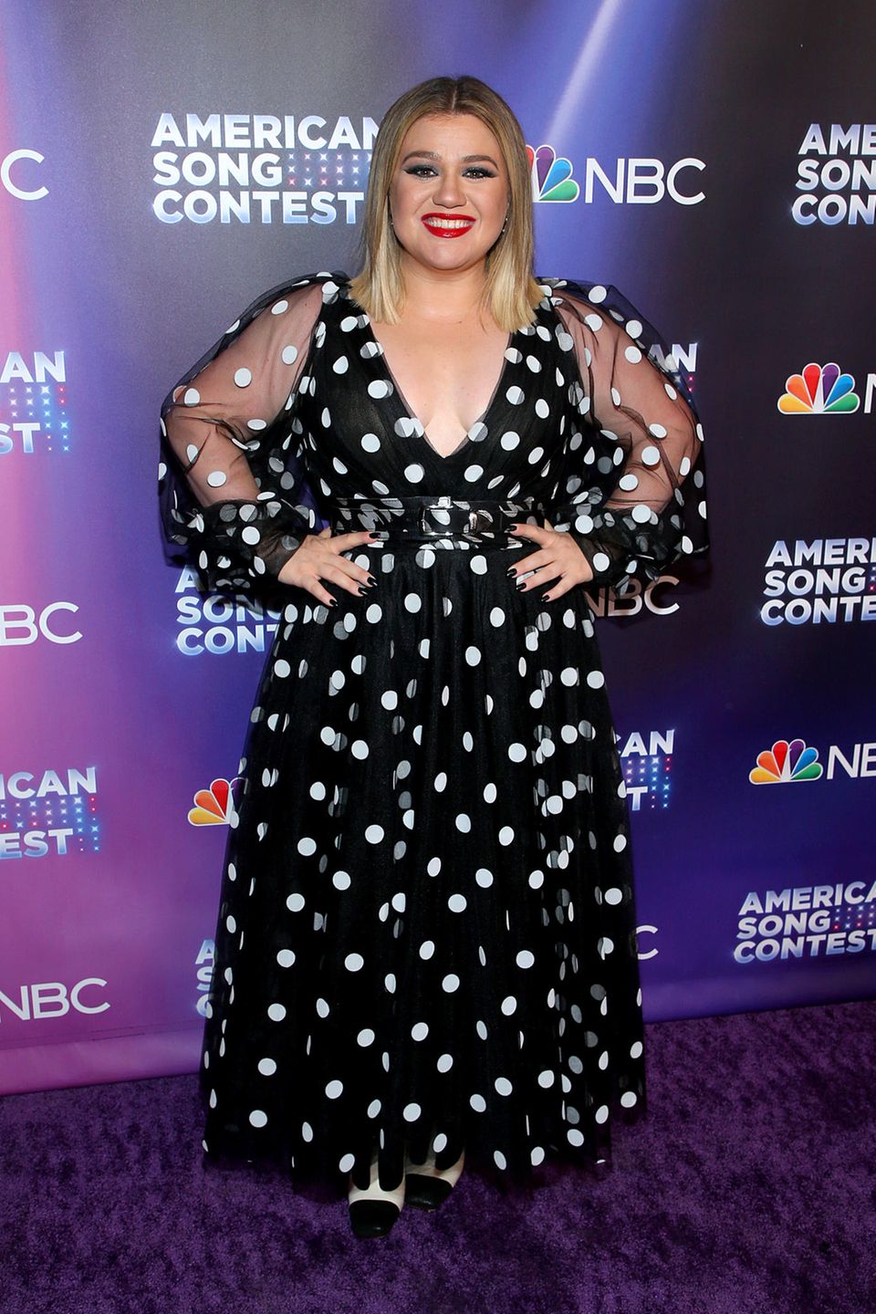 Sängerin Kelly Clarkson verzaubert auf dem Red Carpet der Premiere von "American Song Contest"  mit einem tief ausgeschnittenen schwarzen Kleid mit weißen Punkten. Dazu kombiniert sie weiße High Heels, schwarz lackierte Nägel und kirschroten Lippenstift. Das in A-Linie geschnittene Kleid wird durch die durchsichtigen Blousonärmel zum Hingucker.