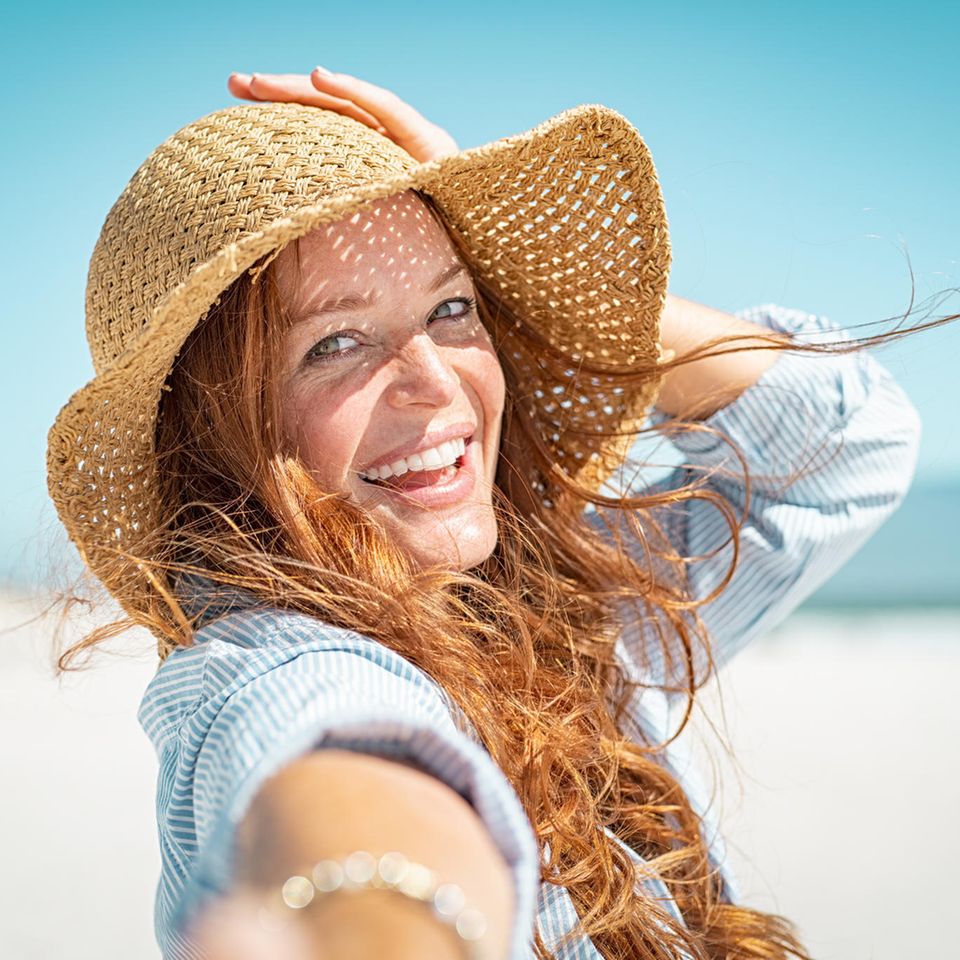 Frau ist am Strand und lacht glücklich in die Kamera während sie ihren Hut festhält.