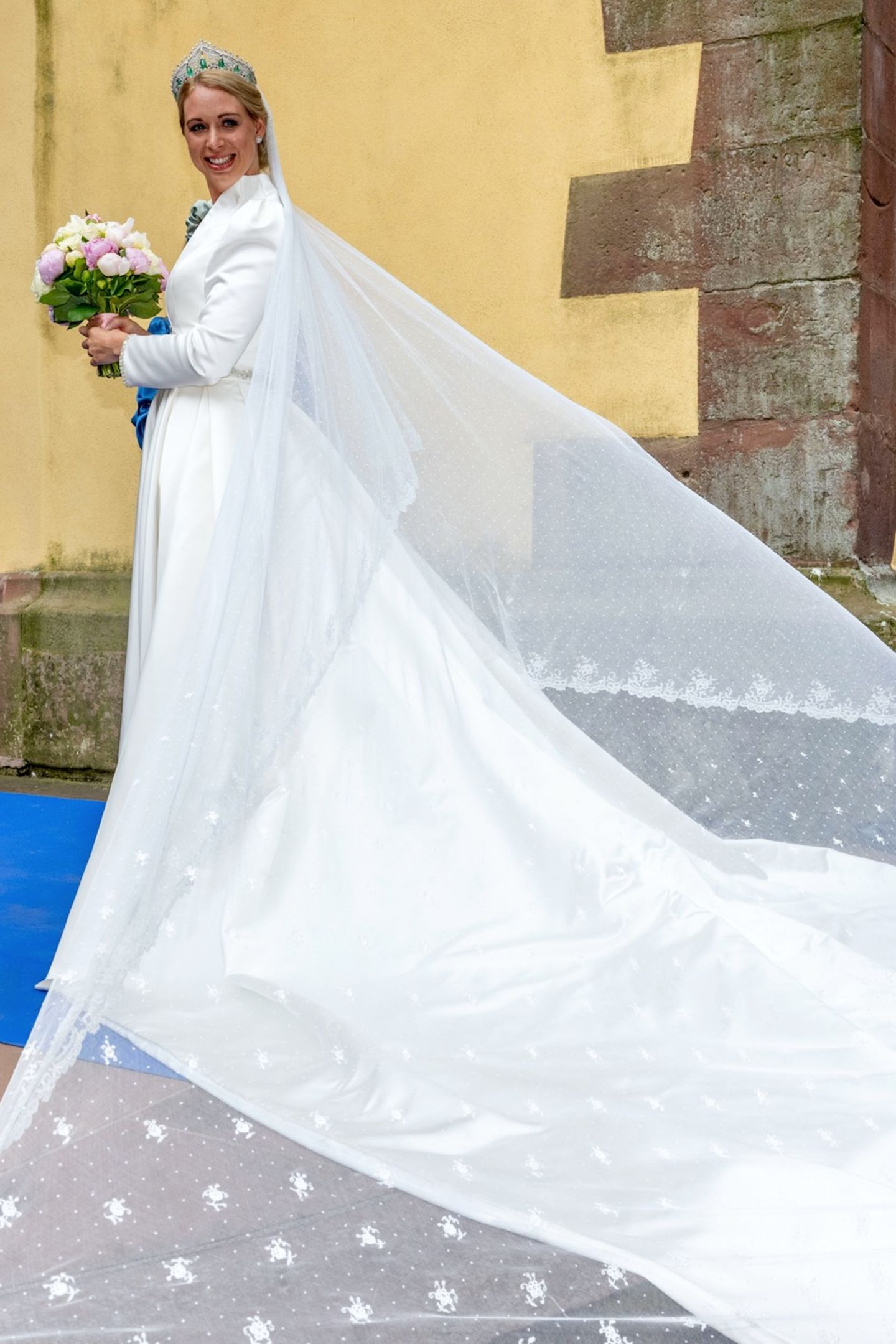Für die Hochzeit mit ihrem Benedikt wählt Prinzessin Amelie zu Löwenstein-Wertheim-Freudenberg ein weißes Brautkleid mit XL-Schleppe. Dazu trägt sie ein edles Diadem mit Smaragden und einen Brautstrauß aus rosafarbenenen Pfingstrosen und weißen Rosen.