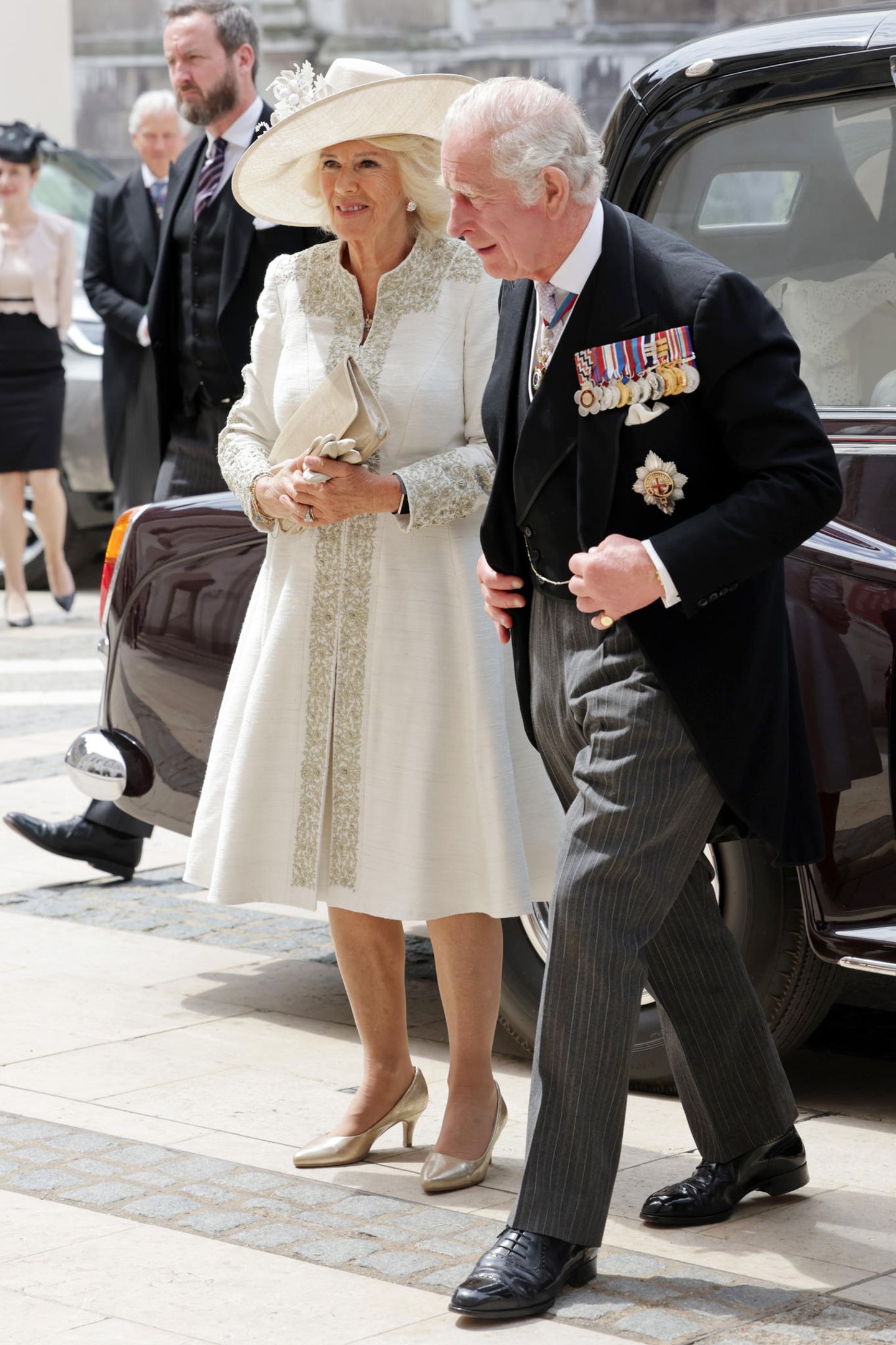 Herzogin Camilla zeigt sich an der Seite von Prinz Charles in einem elfenbeinfarbenen Mantelkleid aus festem Seidenstoff, das an den Säumen kunstvoll verziert ist.