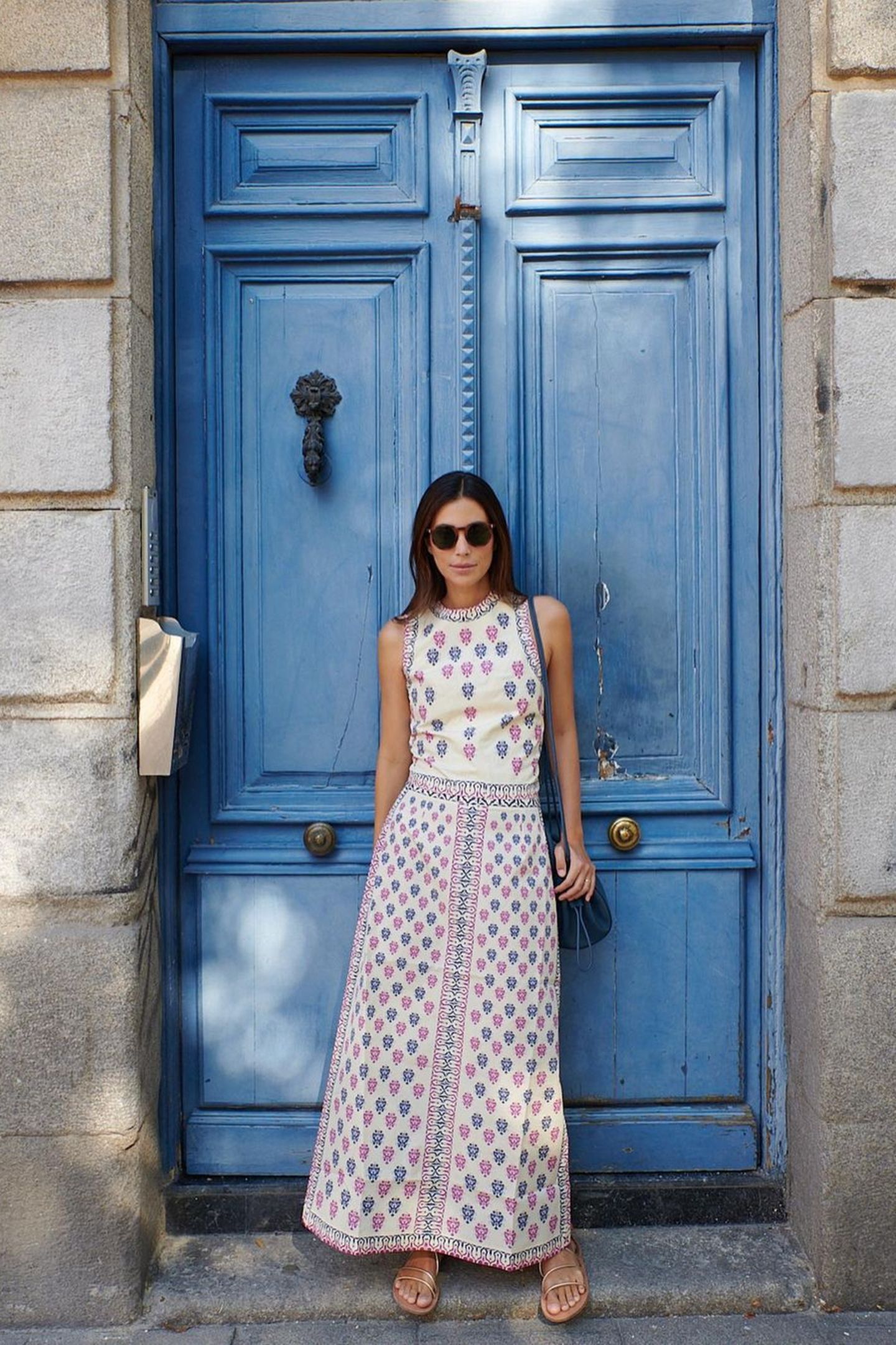 In einem Traum-Kleid posiert Alessandra de Osma vor einem blauen Hauseingang. Der Kontrast zu dem Hintergrund lässt das Kleid noch besser wirken. Das weiße Maxikleid ist verziert in einem abwechselndem Muster aus Pink und Blau. Das einzigartige Outfit kombiniert sie mit schlichten Sandalen und einer dunkelblauen Tasche. Wenn der Look mal nicht nach Urlaub schreit! 