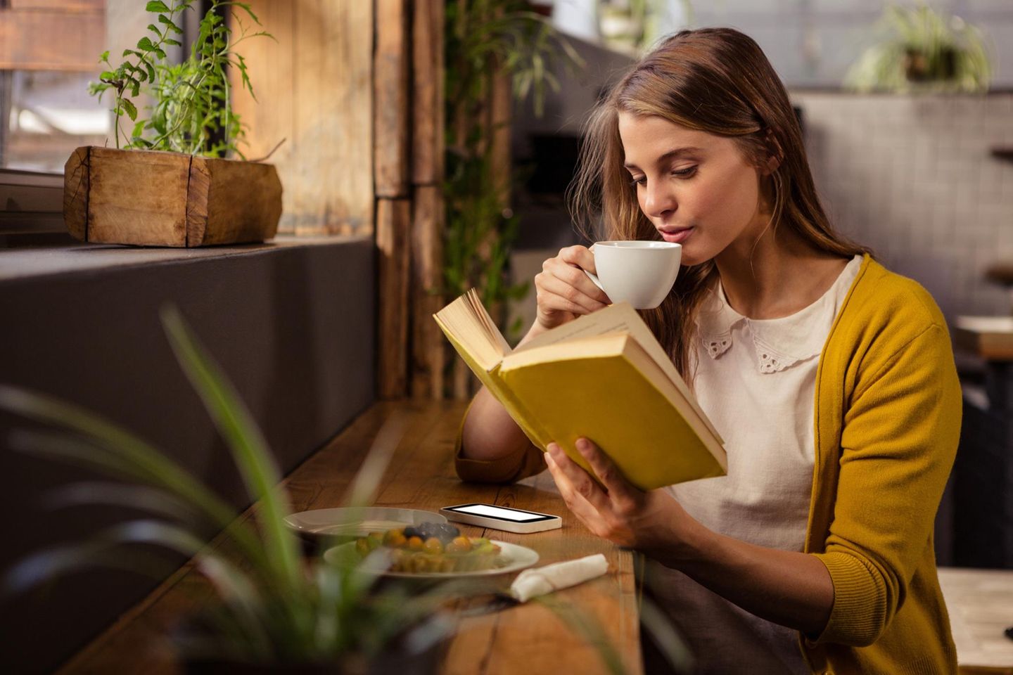 Frau liest ein Buch neben ihrem Smartphone: Diese 7 Trendbücher sind zu Recht Bookstagram-Hypes