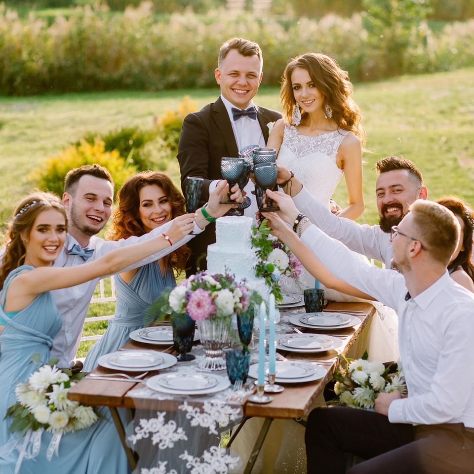 Brautpaar und Gäste in schicken Kleidern an Hochzeits-Tafel
