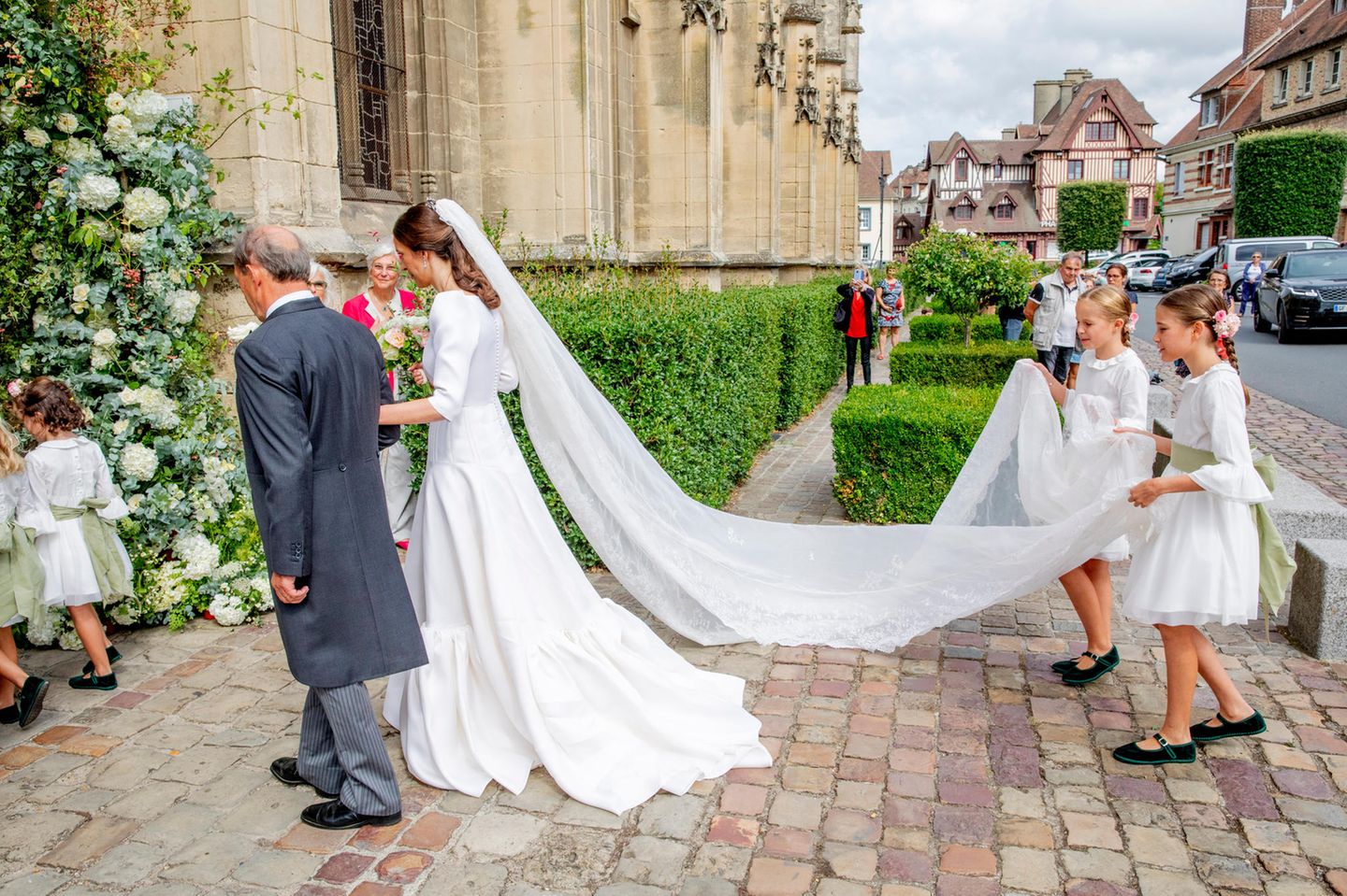 Caroline Philippe trägt ein elegantes mehrstufiges Brautkleid mit einem V-Ausschnitt, langen Ärmeln und einem beeindruckend verzierten Schleier aus feiner Spitze, der von zwei Brautjungfern getragen wird – reduziert, aber wunderschön. 