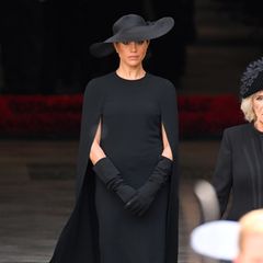 Herzogin Meghan trifft bei der Beerdigung von Queen Elizabeth ein. Die Frau von Prinz Harry trägt ein Cape-Dress von Stella McCartney in Midilänge, dazu eine schwarze Strumpfhose sowie einen schwarzen Hut von Dior. Um ihre Arme zu bedecken, greift Meghan in der Kirche zu langen schwarzen Handschuhen.