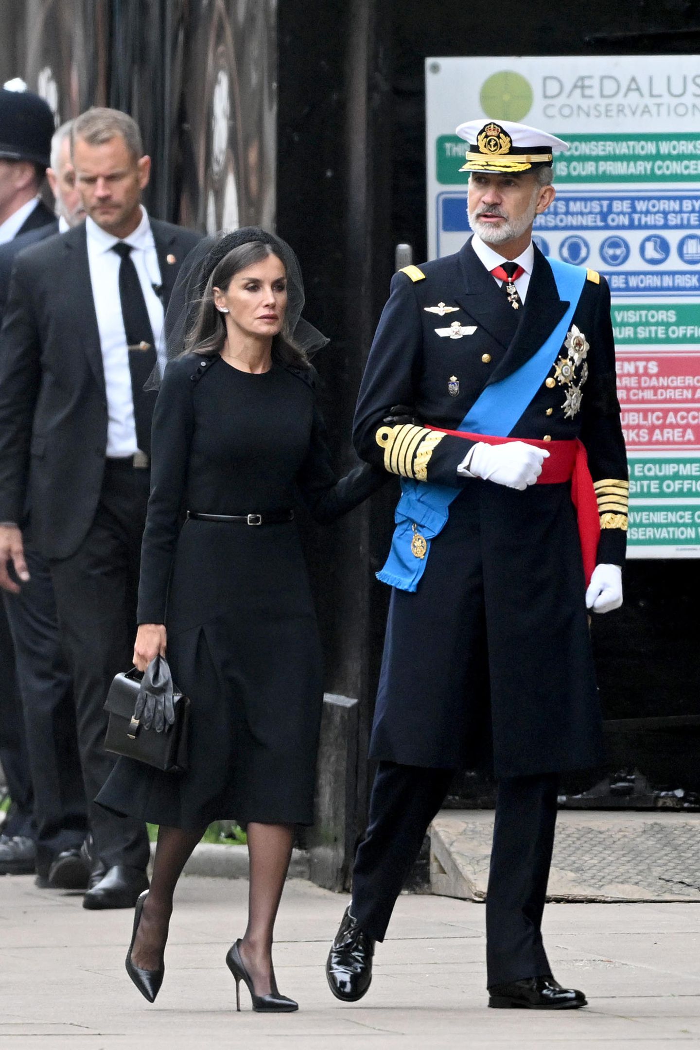 Königin Letizia von Spanien wählt ein schwarzes Kleid mit Taillengürtel von Carolina Herrera. An ihrem schwarzen Haarreif steckt ein Netzschleier. In der Hand hält sie neben ihren schwarzen Lederhandschuhen auch eine Handtasche von Giorgio Armani. 