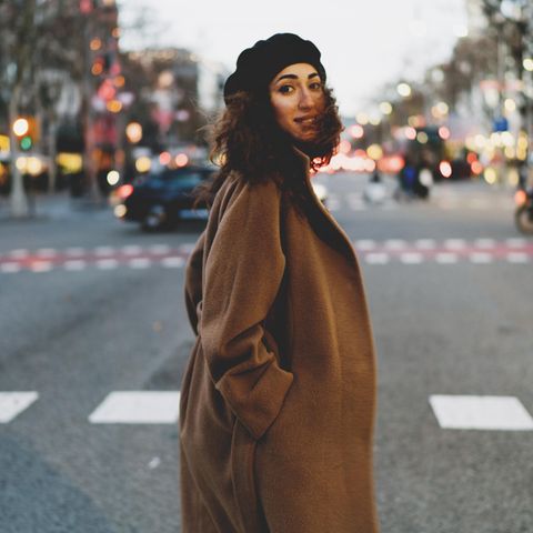 Frau steht auf einer großen Straße und trägt einen braunen Wintermantel