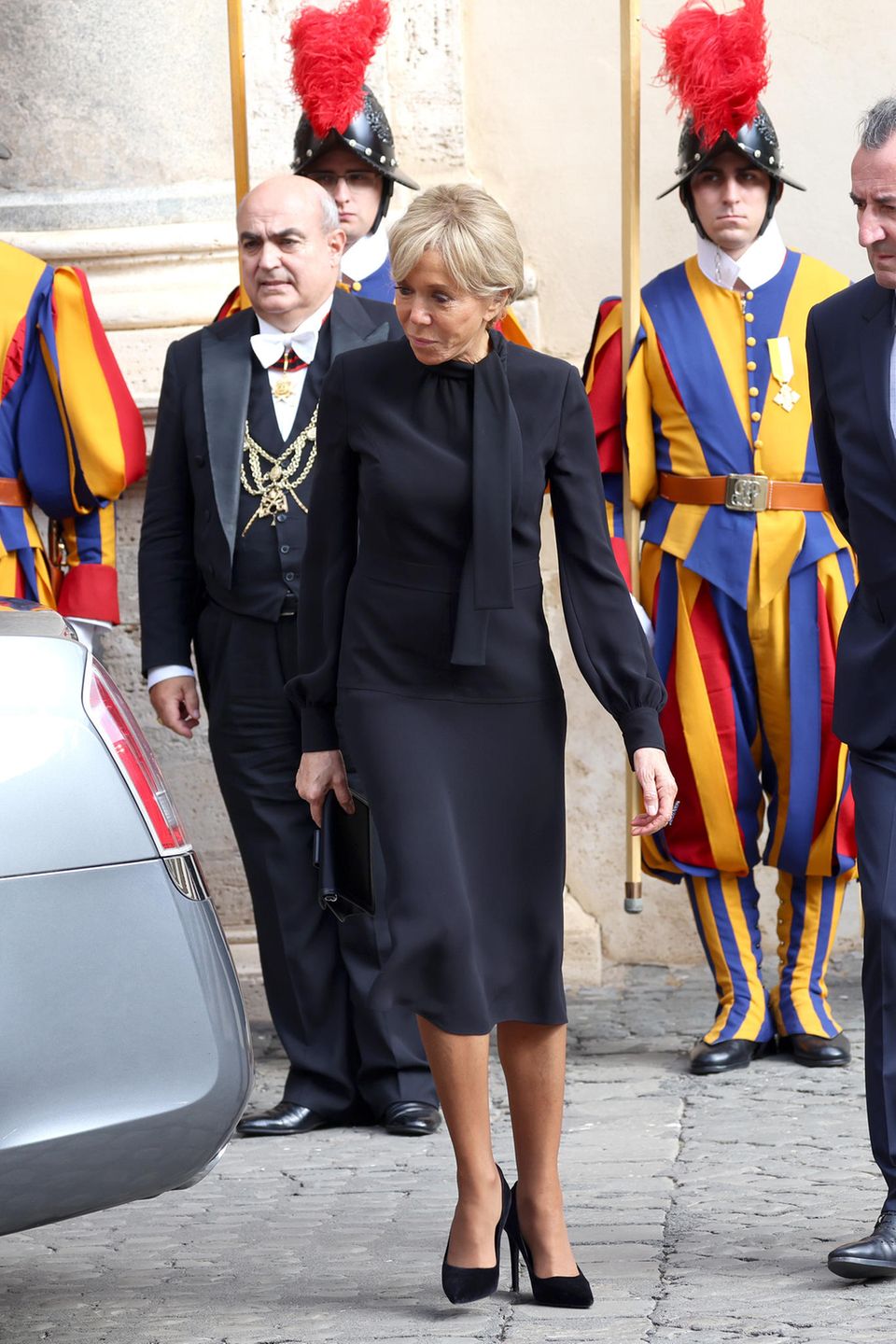 Brigitte Macron begleitet ihren Gatten, Präsident Emmanuel Macron, zu einer privaten Audienz bei Papst Franziskus im Vatikan. Dem Anlass angemessen, zeigt sich die Première Dame in einem schwarzen Kleid in Midi-Länge. Dabei ist das Kleid durch den leichten, fließenden Stoff und den gebundenen Schluppenkragen alles andere als langweilig. Die Clutch von Louis Vuitton und Pumps passt sie farblich an das Kleid an, während sie das Haar seriös zum Dutt gebunden trägt.