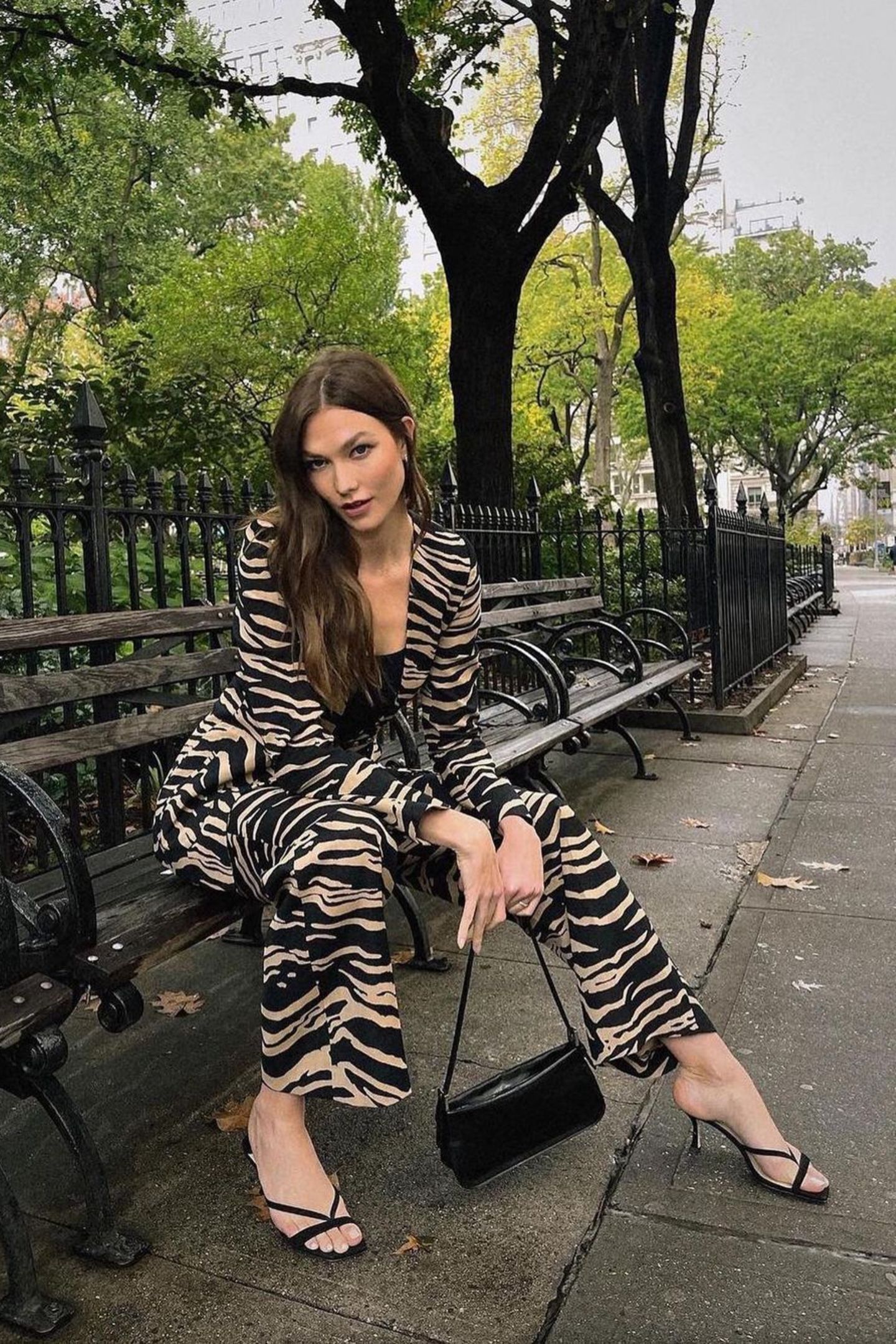 Auf Safari im Central Park! Karlie Kloss posiert auf einer Bank in einem sexy Anzug mit Zebra-Print in Braun-Schwarz. Ihre Accessoires hält sie dezent und kombiniert dazu zarte Sandaletten von Jimmy Choo und eine Baguette-Tasche, ebenfalls in Schwarz. Die Haare des Models fallen in natürlichen Wellen über die Schulter. 