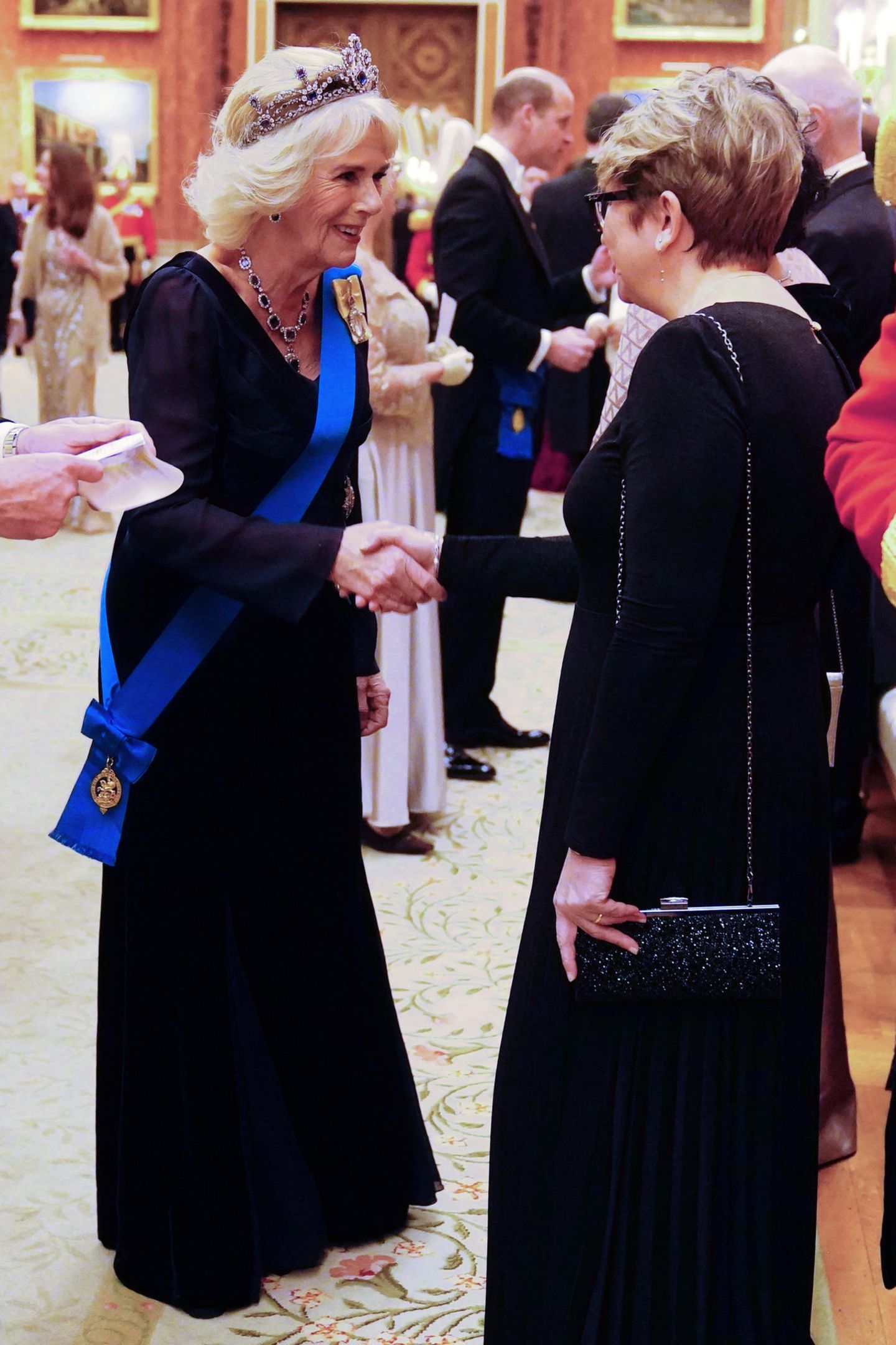 Unter dem besonderen Mantel trägt Camilla ein Kleid von Anna Valentine. Das dunkelblaue bodenlange Dress kommt mit einem V-Ausschnitt daher und gibt so den Blick auf ganz besondere Juwelen, die Camilla zu Ehren von Queen Elizabeth trägt, frei.