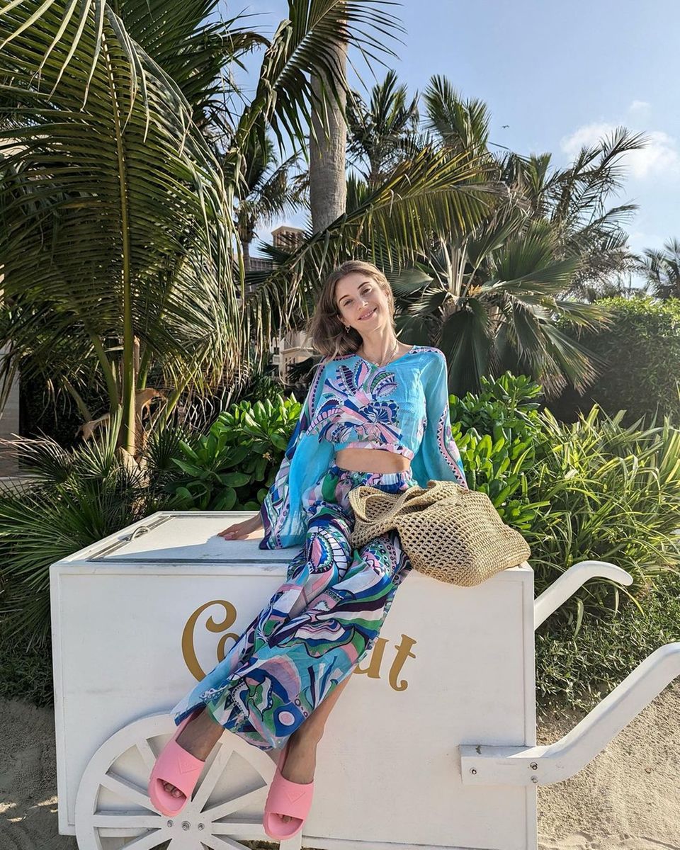 Palmen, Strand und ein breites Grinsen. Lachend posiert Cathy Hummels in einem blauen Zweiteiler, welcher verschiedene, bunte Mandala-Muster ziert. Die rosafarbenen Badeschuhe und die geflochtene Tasche vervollständigen das Ensemble aus Maxirock und Bluse und beweisen, wie einfach sich ein luftig-leichter Strandlook gestalten lässt.
