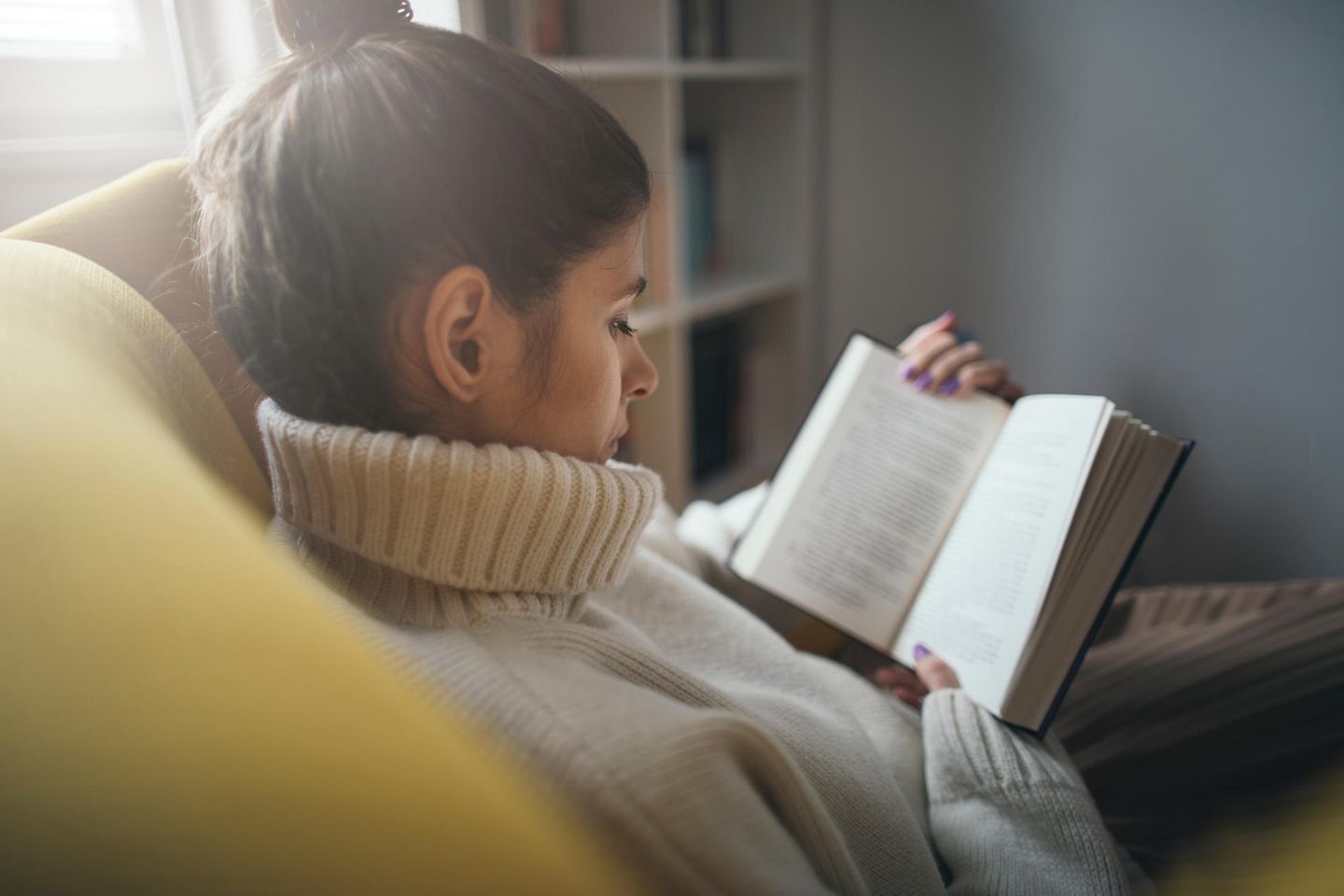 Frau liest im Sessel: Diese 5 Bücher sind perfekt, um dem Winterblues zu entkommen