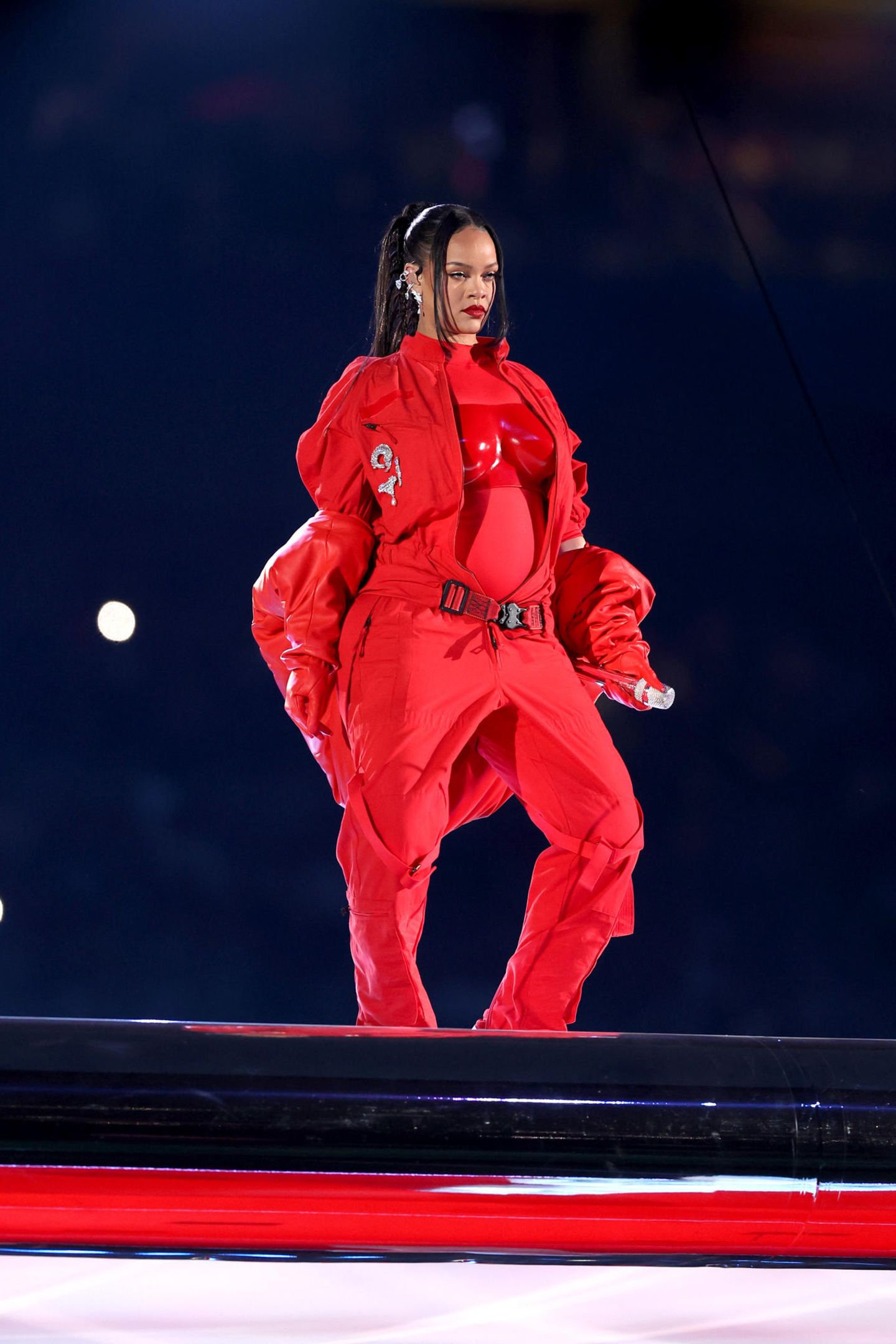 Sie macht Rot zu ihrer Farbe: Sängerin Rihanna trägt für ein Medley ihrer besten Hits zur Halbzeitshow des Super Bowls einen Einteiler von Loewe, worüber sich das passende Bustier verbirgt. Der Gürtel, der leicht über ihrer Hüfte sitzt, verdeutlicht ihre freudigen Neuigkeiten noch einmal stärker: Rihanna erwartet ihr zweites Baby. Aber auch die kurze Daunenjacke von Alaïa verleiht ihren Baby-News den richtigen Rahmen. Motto: Freie Sicht auf den Bauch! Doch ...