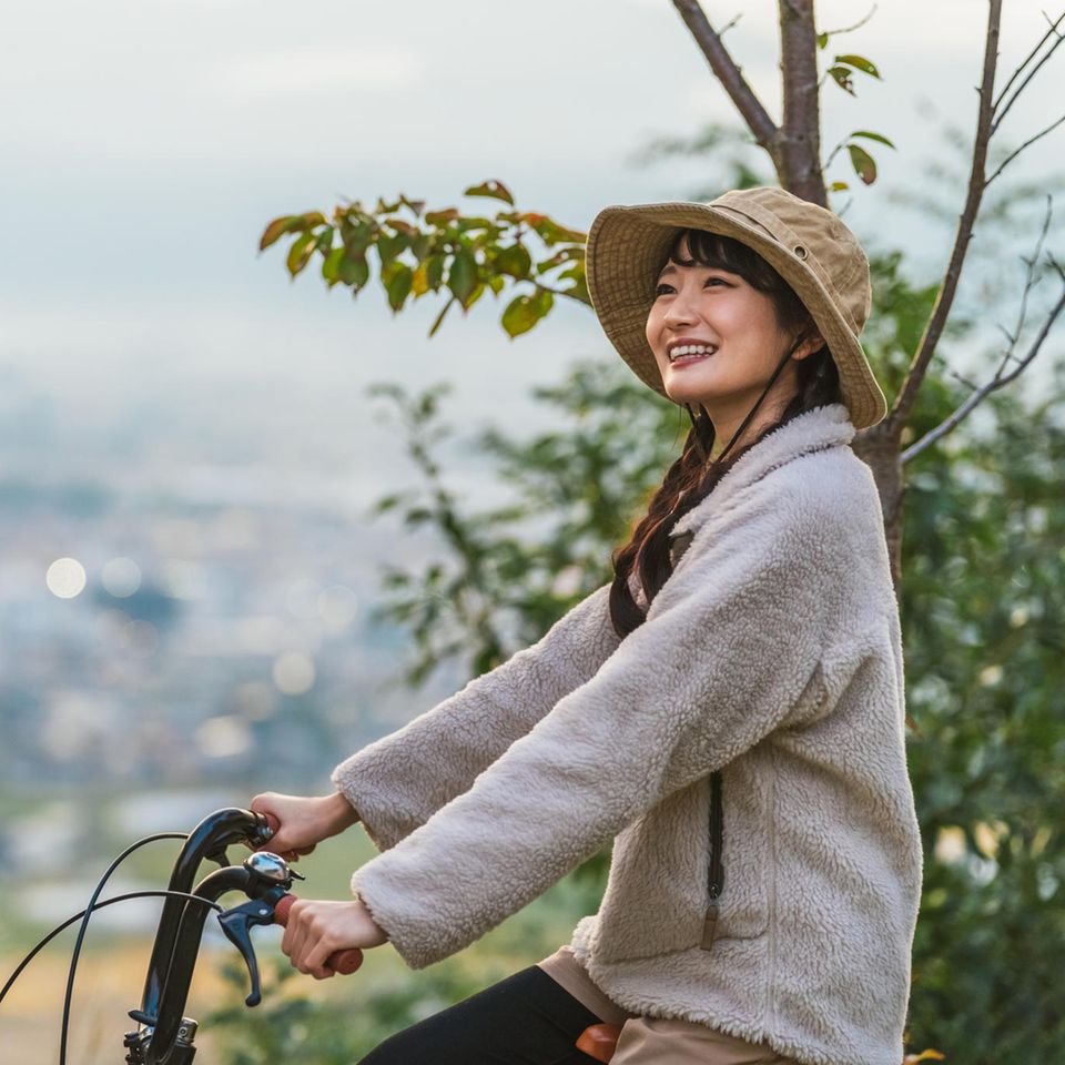 Die perfekte Fleece-Jacke: Warum jetzt alle auf das Trend-Material setzen, junge Frau auf Fahrrad mit Hut und Fleece-Jacke