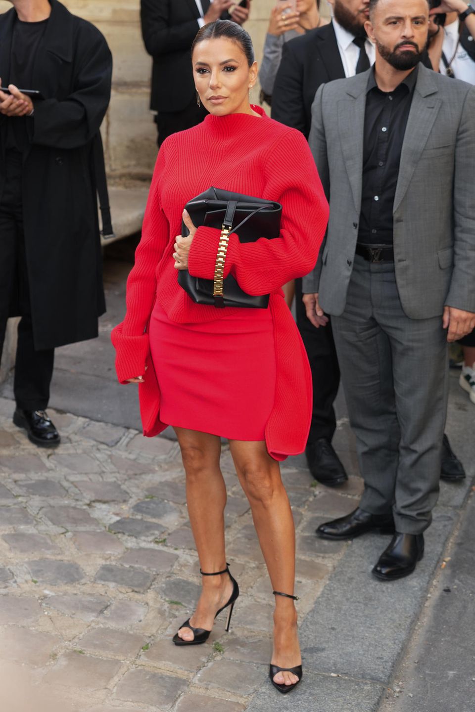 Eva Longoria setzt für die Victoria Beckham Fashion Show auf eine Kombination aus eng anliegendem Rock und Pullover mit länger geschnittenen Ärmeln. Zum Rot der beiden Teile kombiniert sie Schwarz: Ihre Riemchen-Sandalen und Tasche vollenden die klassische Farbkombination. 