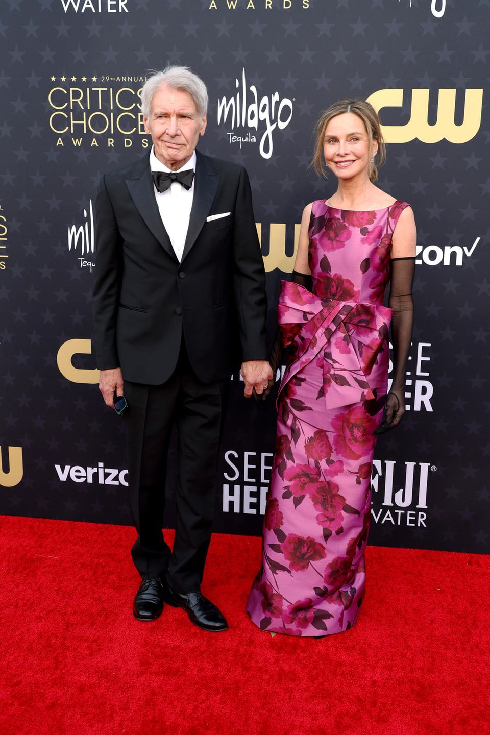 Harrison Ford und Ehefrau Calista Flockhart haben auf dem roten Teppich ihren Holly Golightly und Paul Varjak-Moment. Sie bezaubert in einem von Frühstück bei Tiffanys inspiriertem Schleifenlook in Pink, er in klassischem schwarzen Tuxedo. 
