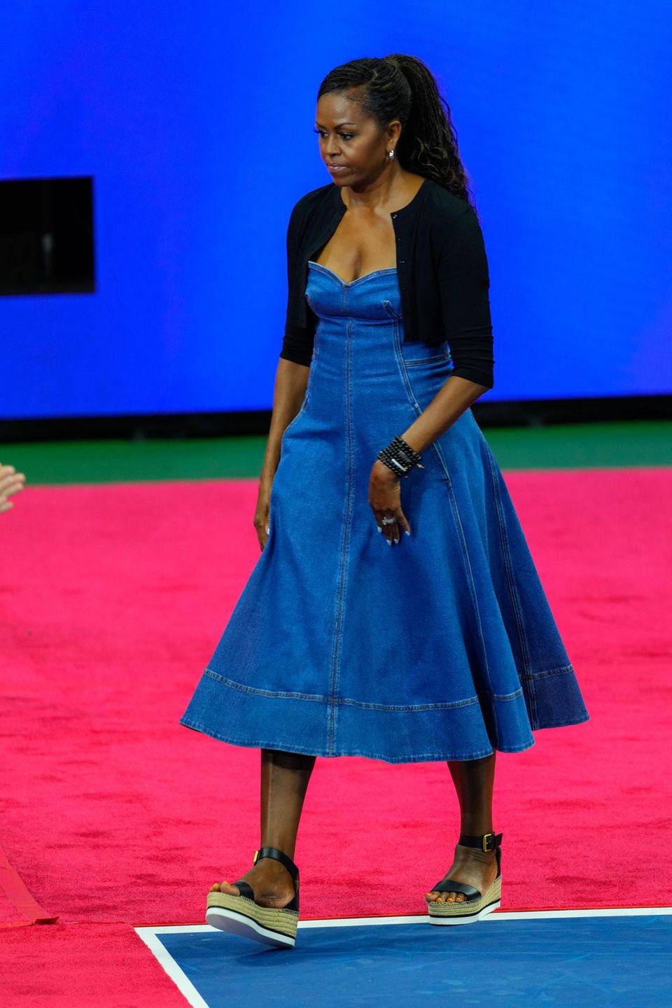In einem lässigen, aber doch eleganten Look besucht Michelle Obama 2023 die "US Open Tennis Championships" in New York. Ihr Stilgefühl stellt sie in einem Midi-Jeanskleid mit einem relativ ausgestellten Rock unter Beweis. Dazu kombiniert sie ein schwarzes Bolero-Jäckchen und Sandalen mit Plateausohlen.